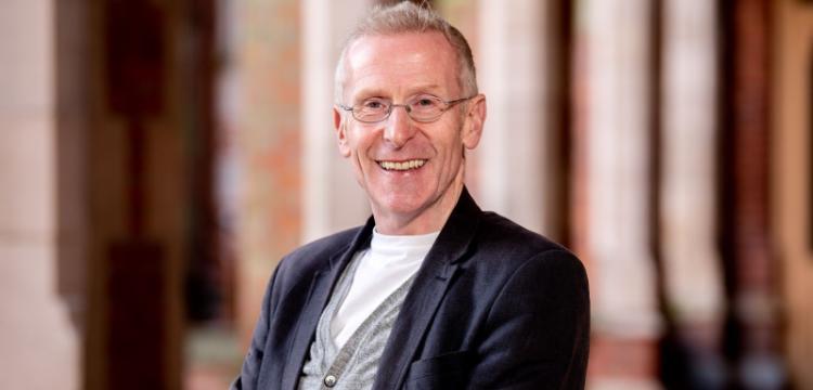 head and shoulders photo of a smiling man with short grey hair and wire-rimmed glasses, wearing a white t-shirt, grey cardigan and navy jacket